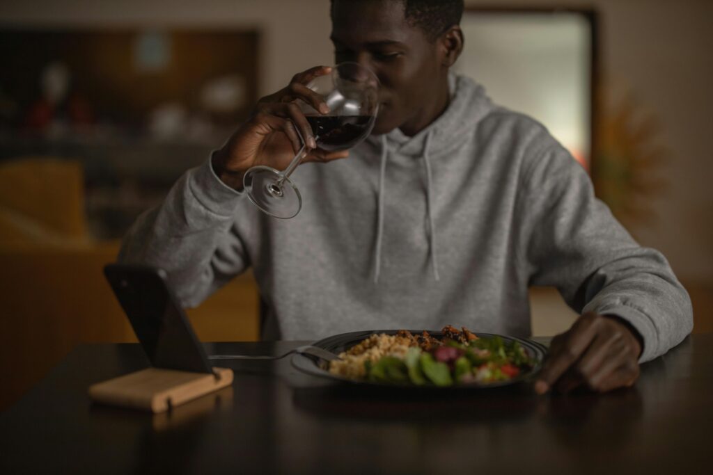 man in a gray hoodie drinking wine and eating dinner.